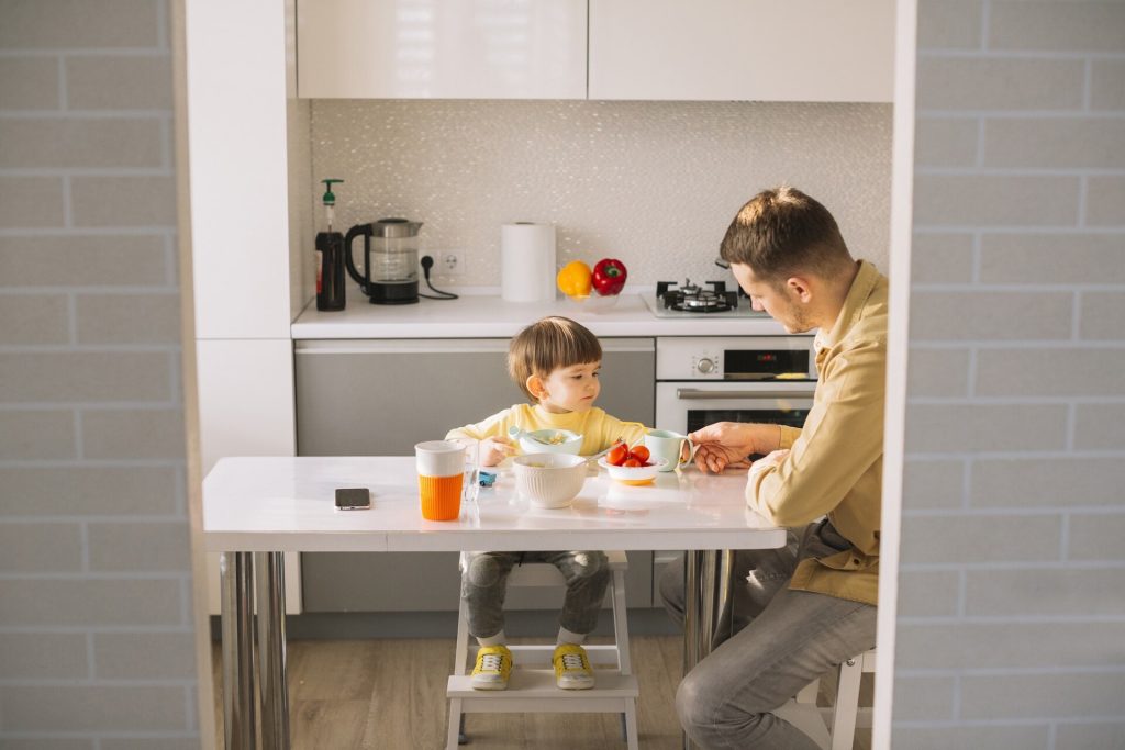Litet barn i en hög barnstol av trä vid ett vitt köksbord med sin pappa
